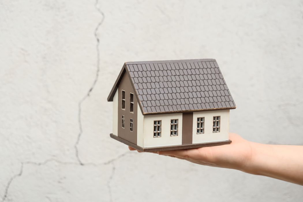 Maqueta de casa de dues plantes sobre una mà, amb fons de paret grisa esquerdada. Concepte de terratrèmol