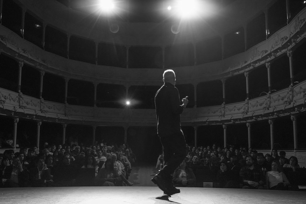 Escenario de teatro con persona de espaldas bajo focos, frente a público en platea y balcones, interior clásico circular con iluminación dramática en blanco y negro.