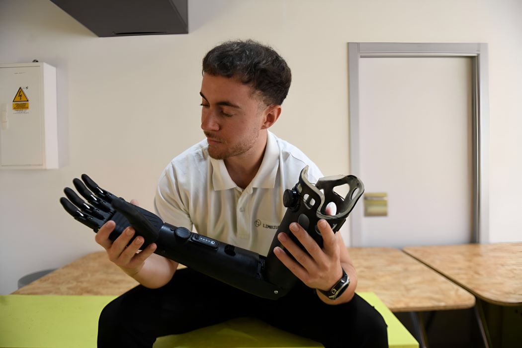 Persona sentada sostiene y observa un brazo protésico robótico desmontado en un entorno interior, destacando innovación tecnológica y desarrollo de prótesis