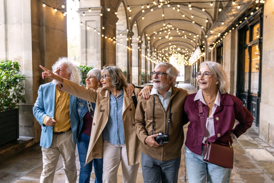 Grupo de personas mayores paseando juntas por una galería con arcos e iluminación cálida; una persona señala al frente mientras otra lleva una cámara