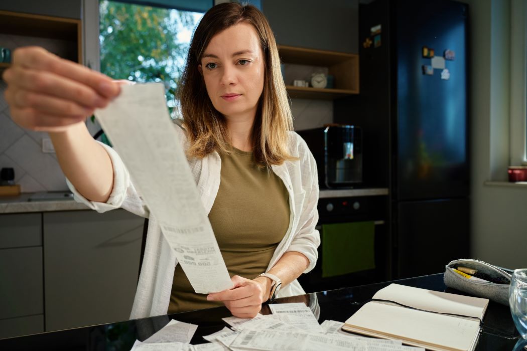 Persona en cocina revisando un recibo largo con varios tickets y una libreta sobre la encimera, en un entorno doméstico moderno.
