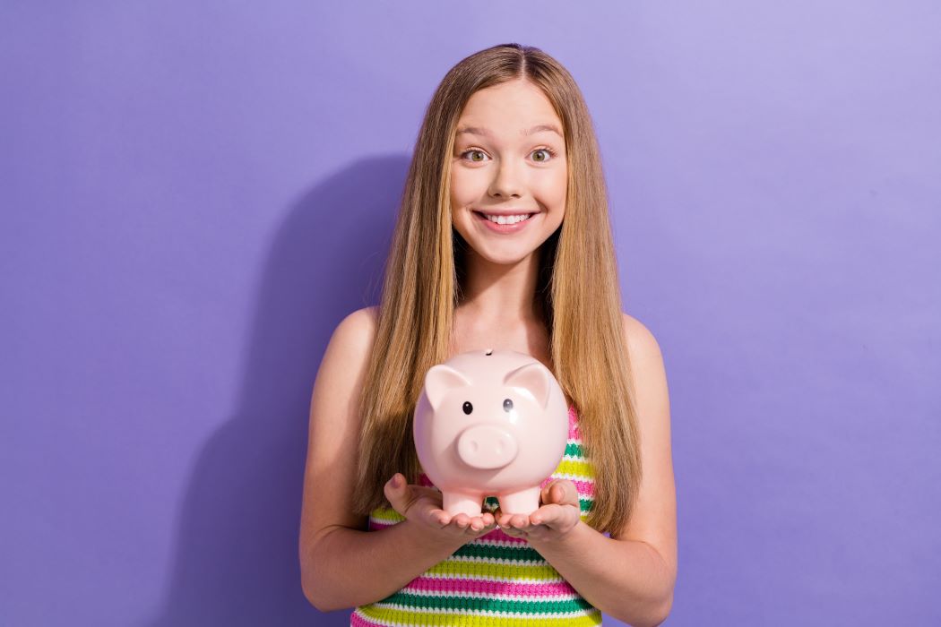 Adolescente de pelo largo sosteniendo una hucha de cerdito rosa frente a un fondo lila uniforme.