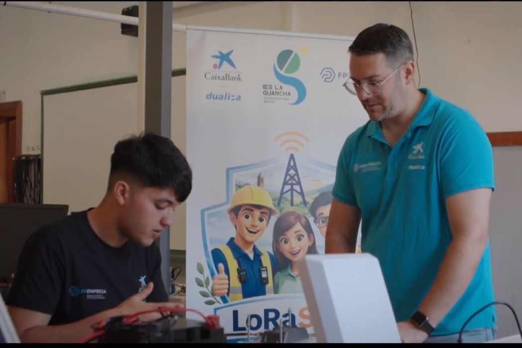 Alumne d'FP i docent revisen un projecte tecnològic en una aula, al costat d'un cartell del sistema LoRaSOS desenvolupat per millorar la seguretat de les persones grans