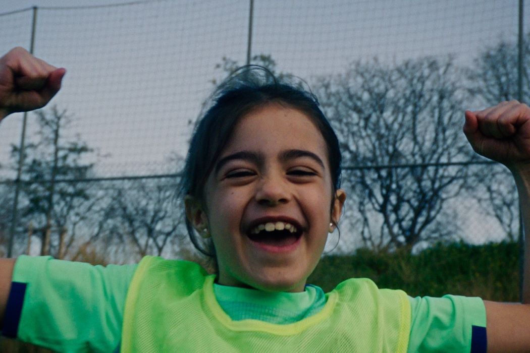 Niña jugando al fútbol en un campo deportivo, con peto verde fluorescente y los brazos levantados frente a una red de protección
