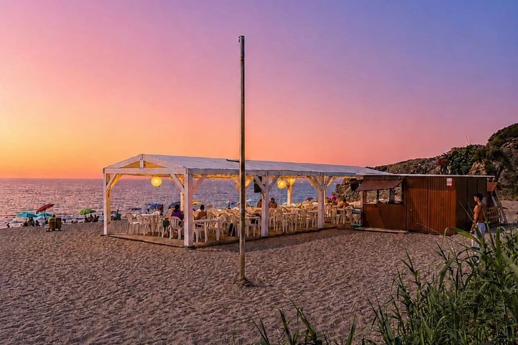 Chiringuito de playa sobre la arena al atardecer, con terraza cubierta, mesas y sillas junto al mar, bajo un cielo de tonos rosados y anaranjados 