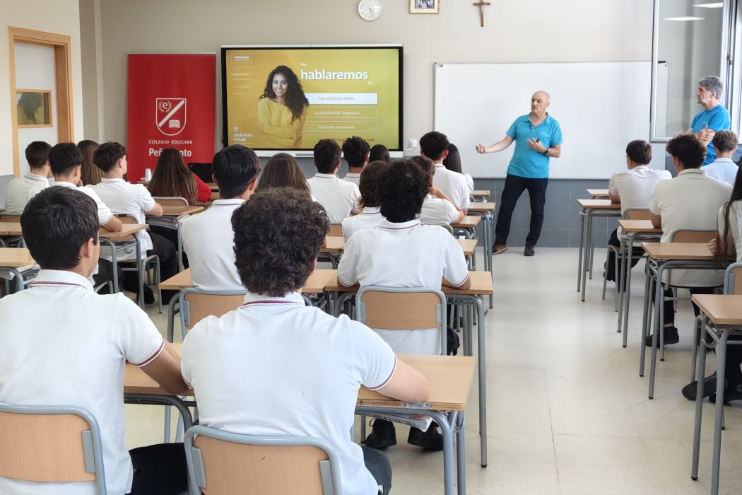 Voluntari sènior imparteix una xerrada a estudiants en una aula, al costat d’una pantalla de presentació i una pissarra.