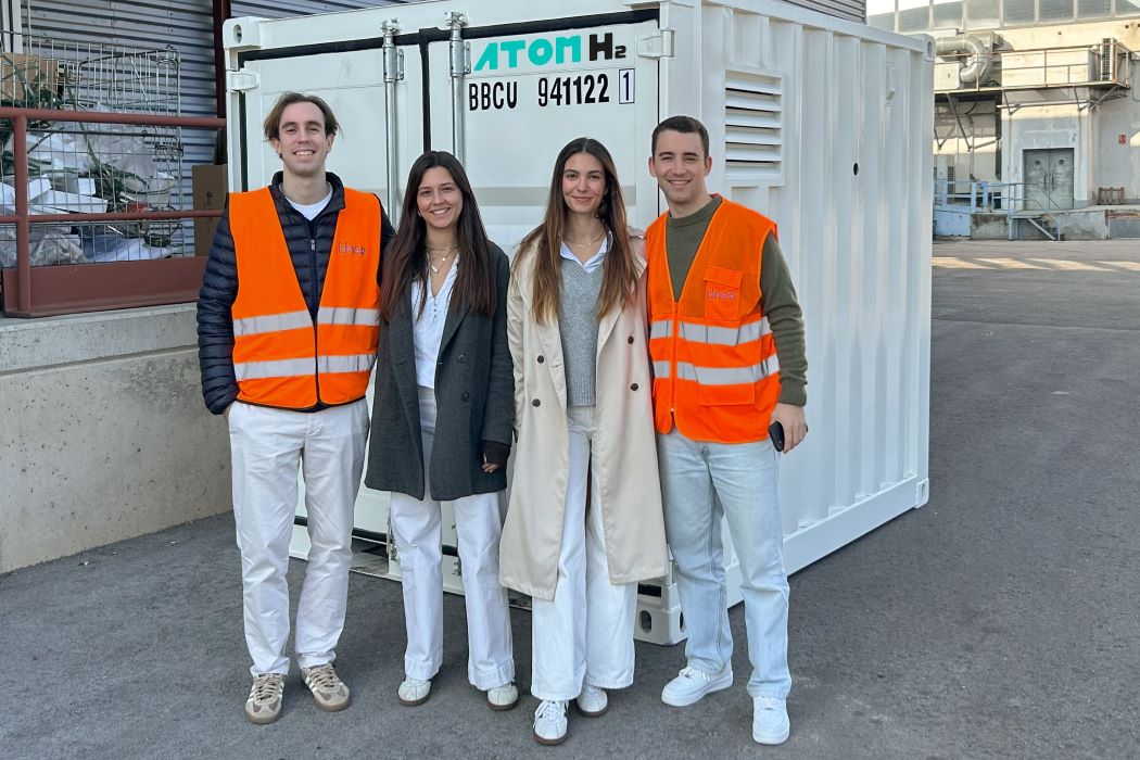 Cuatro personas posan frente a un contenedor blanco de hidrógeno con el logotipo “ATOM H2” en una zona industrial; dos llevan chalecos reflectantes naranjas
