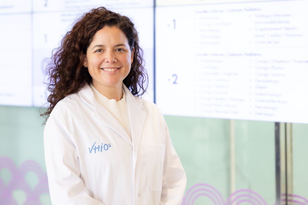 Elena Garralda con bata blanca en primer plano, frente a una pantalla con información científica desenfocada en un entorno de laboratorio o investigación