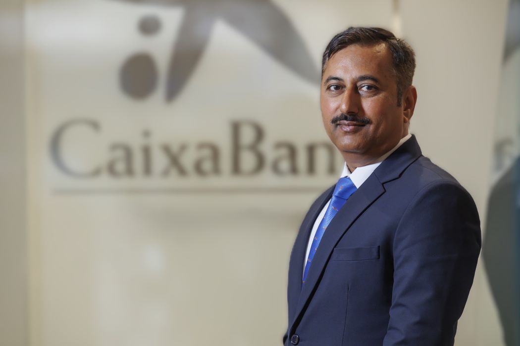 Rahul Prakash amb vestit blau marí i corbata blava posant en una oficina, amb el logotip de CaixaBank desenfocat al fons