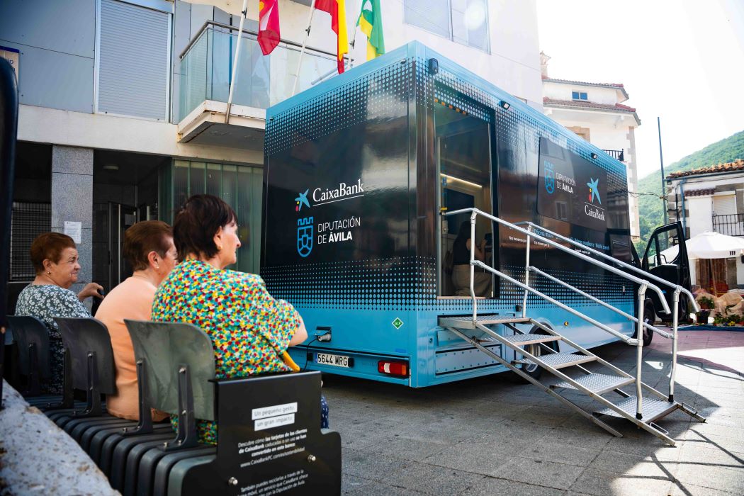 Oficina mòbil de CaixaBank estacionada a Serranillos (Àvila), davant d’un grup de veïnes assegudes en bancs fabricats amb targetes reciclades.