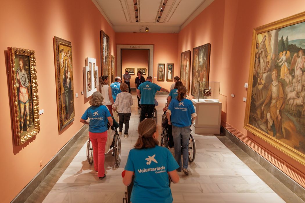 Grup de voluntariat acompanya persones usuàries de cadira de rodes durant una visita accessible a una sala de museu amb pintures exposades
