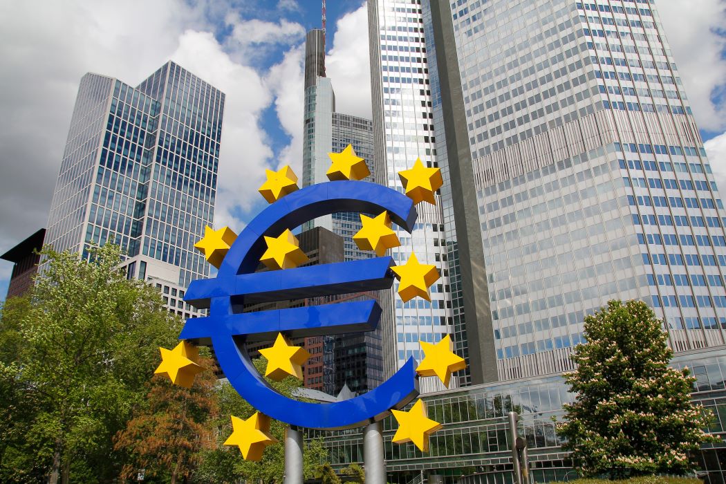 Símbolo del euro rodeado por estrellas amarillas, situado en una plaza urbana con rascacielos modernos al fondo.