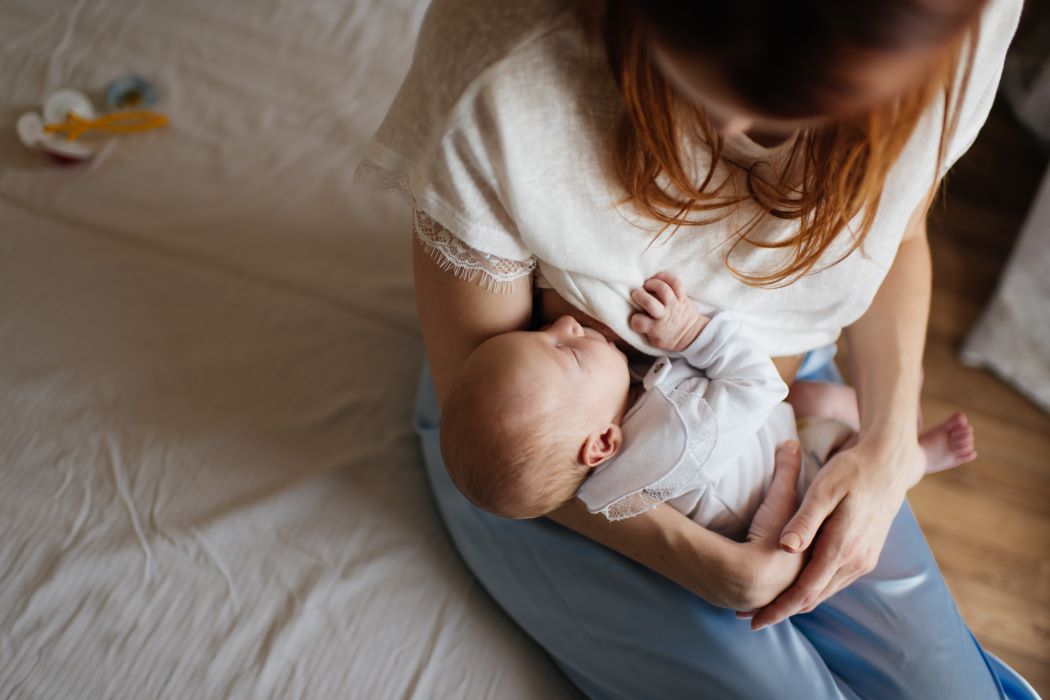 Una dona persona en un llit sosté en braços un nadó mentre l'alimenta. Vista zenital en un entorn domèstic amb roba clara i llum natural