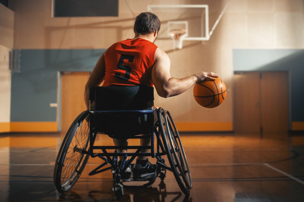 Un hombre en silla de ruedas lanza un balón de baloncesto hacia una canasta mientras una mujer espera recibirlo