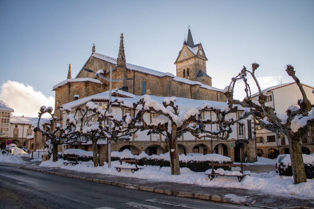 Església de pedra i plaça nevada en un poble de muntanya, amb arbres podats coberts de neu i bancs davant de l'edifici històric. Espinosa de los Monteros (Burgos)