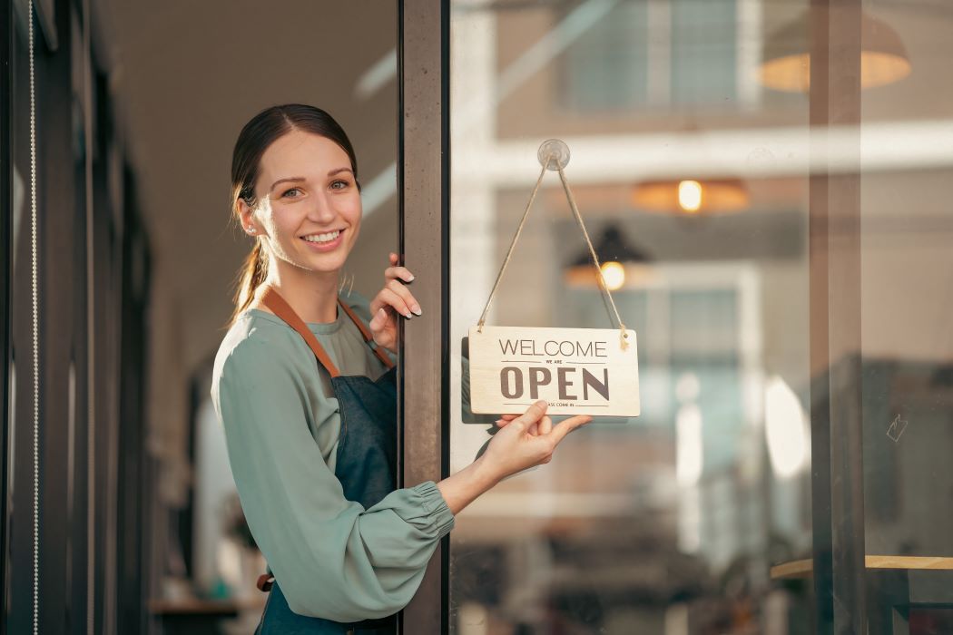 Emprendedora en la entrada de un comercio sosteniendo un cartel de “Open” en la puerta de cristal, con el establecimiento abierto y visible al fondo