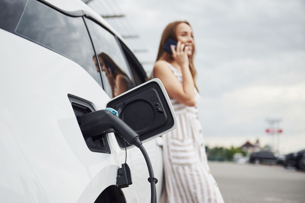 Vehículo eléctrico conectado a un punto de recarga mientras una persona utiliza un teléfono móvil en el exterior, representando la movilidad sostenible