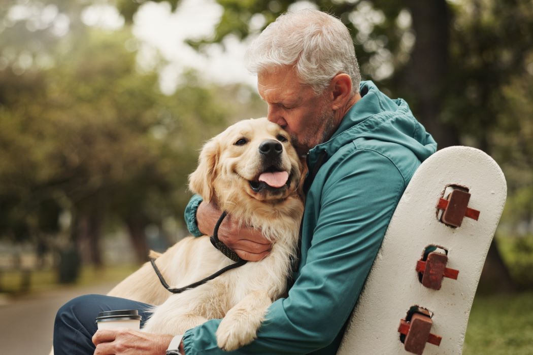 Persona sentada en un banco de un parque abrazando a un perro golden retriever con correa; sostiene un vaso mientras ambos están al aire libre rodeados de árboles
