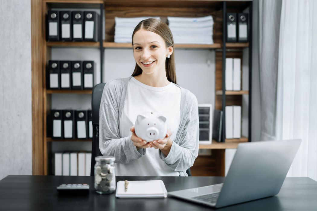 Persona sentada en una oficina mostrando una hucha en forma de cerdo sobre el escritorio, con un portátil, una calculadora y un bote con monedas, símbolo de ahorro financiero.