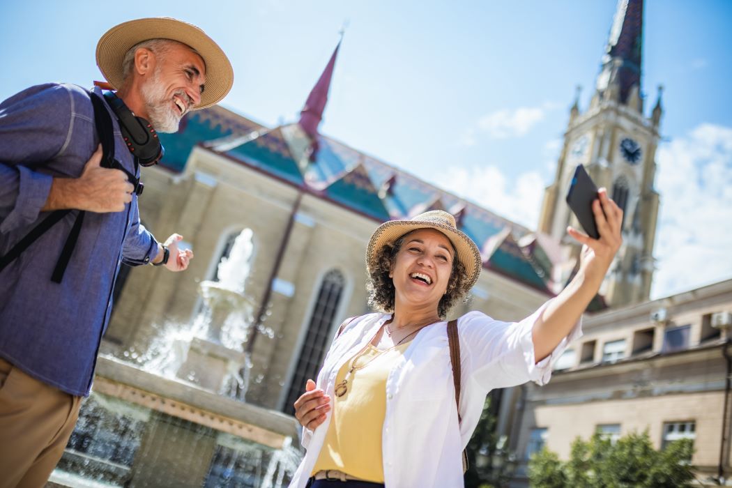 Persona fent una foto amb el mòbil al costat d’una font en una plaça històrica, acompanyada per una altra persona, amb un edifici amb torre al fons