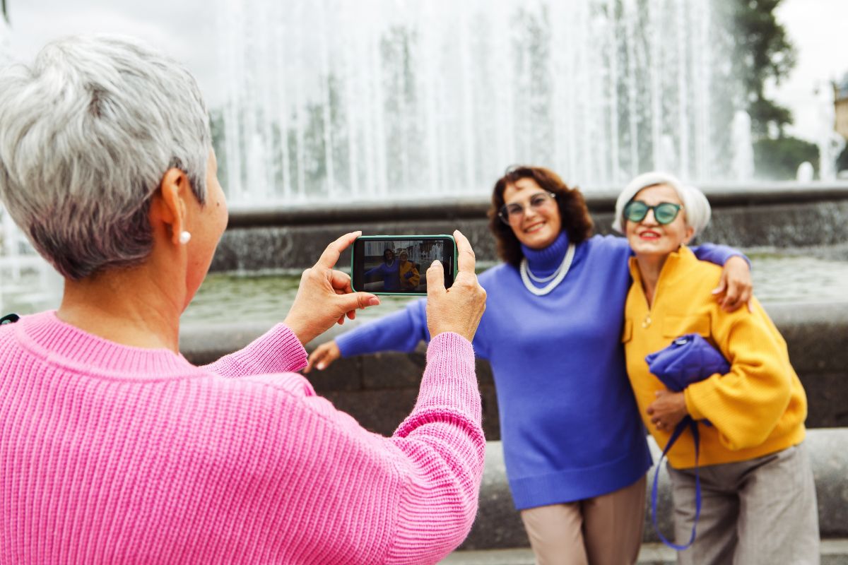 Tres amigues jubilades, alegres i amb roba de colors, es fan una fotografia amb un telèfon i riuen mentre gaudeixen d’un viatge de l’Imserso en una ciutat a l’estiu.