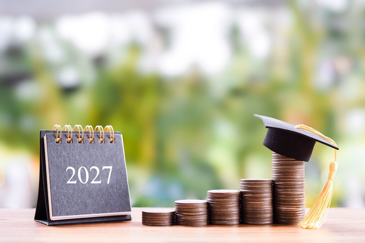 Calendario de 2027 junto a pilas crecientes de monedas, coronadas por un birrete de graduación.