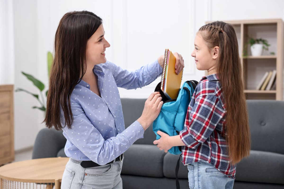 Persona ajudant una nena a preparar una motxilla escolar amb quaderns i material per a la tornada a l'escola a casa.