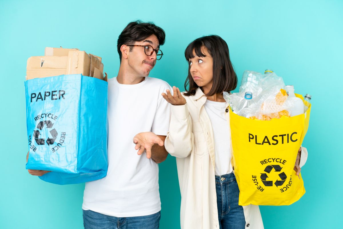 Dos personas sostienen bolsas de reciclaje, una con cartón y papel y otra con envases de plástico, frente a un fondo azul, mostrando la separación de residuos.