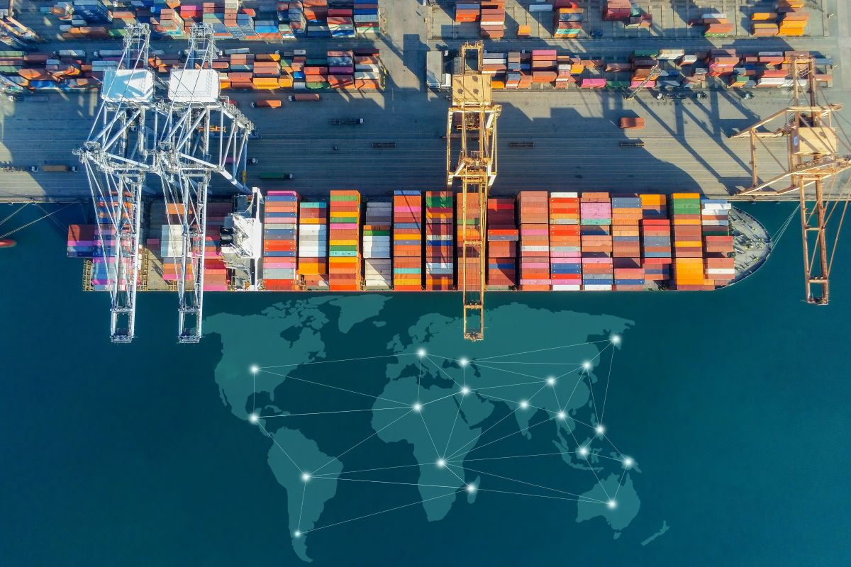Vista aèria d’un port de contenidors amb vaixell de càrrega, grues industrials i un mapa mundial superposat que representa la logística i el comerç internacional