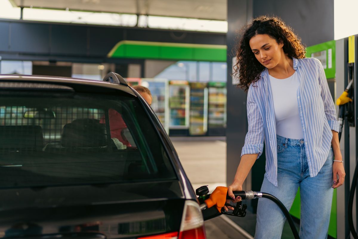 Persona repostando combustible en un vehículo en una gasolinera, con la manguera conectada al depósito y surtidores al fondo