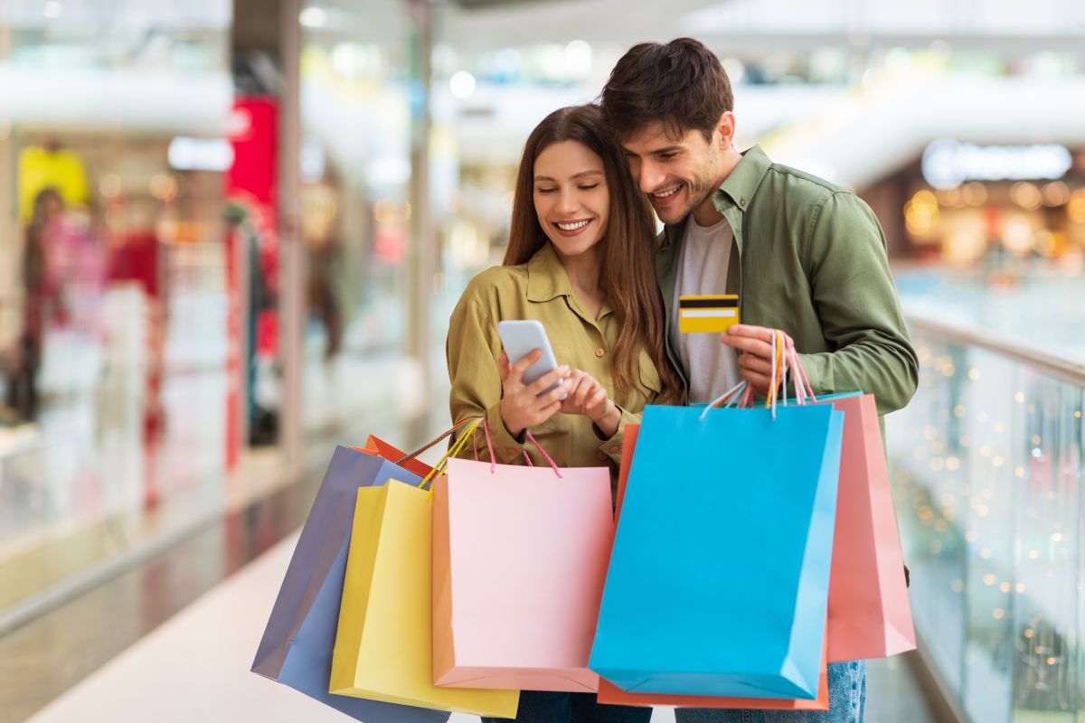 Persones amb bosses de compra consulten un telèfon mòbil en un centre comercial, mentre una d’elles sosté una targeta de pagament
