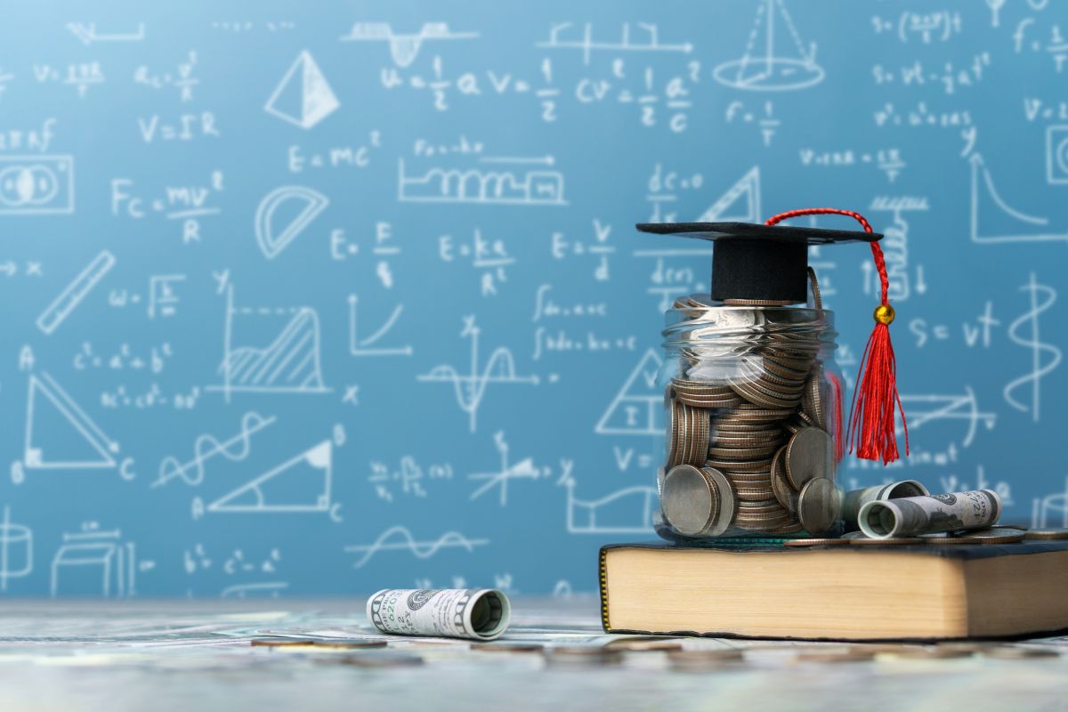 Un pot ple de monedes, amb bitllets al seu voltant i un birret al damunt, reposa sobre un llibre davant d’una pissarra plena de fórmules i diagrames, com a símbol de l’educació financera a l’escola.