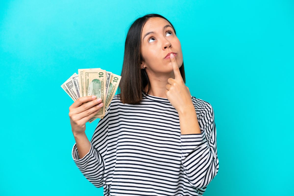 Persona sosteniendo varios billetes de dólar con una mano y apoyando un dedo en la barbilla mientras mira hacia arriba