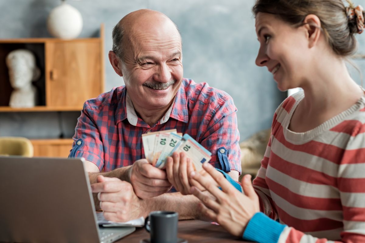 Dues persones assegudes davant un portàtil; una d'elles sosté diversos bitllets d'euro mentre conversen. Concepte de donació de diners entre familiars.