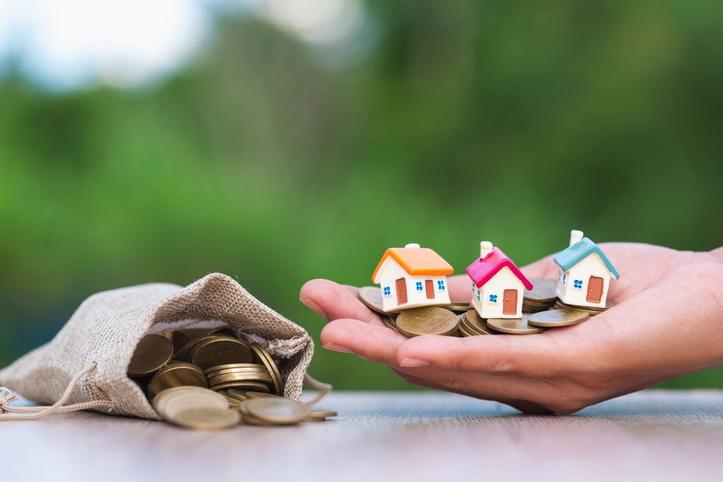 Mano sosteniendo varias casas en miniatura colocadas sobre monedas, junto a una bolsa con monedas, que simboliza el reparto de una herencia