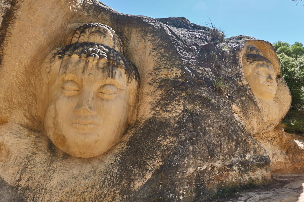 Esculturas de rostros humanos talladas directamente en una gran formación rocosa al aire libre, integradas en el paisaje natural bajo un cielo despejado.