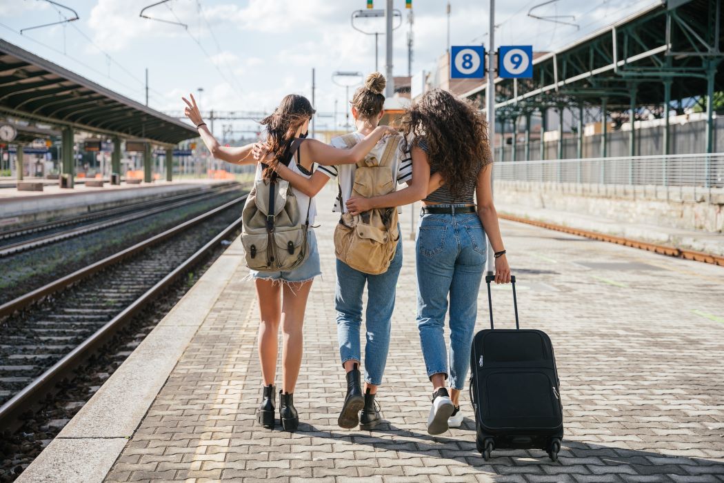Tres mujeres jóvenes con mochilas caminan juntas por un andén de tren, con los brazos sobre los hombros, una de ellas lleva una maleta con ruedas