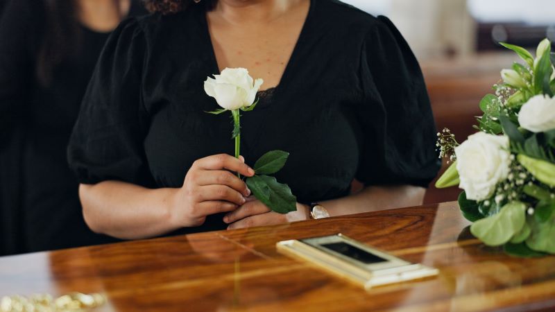 Una mujer vestida de luto sujeta una rosa blanca delante de un f&eacute;retro