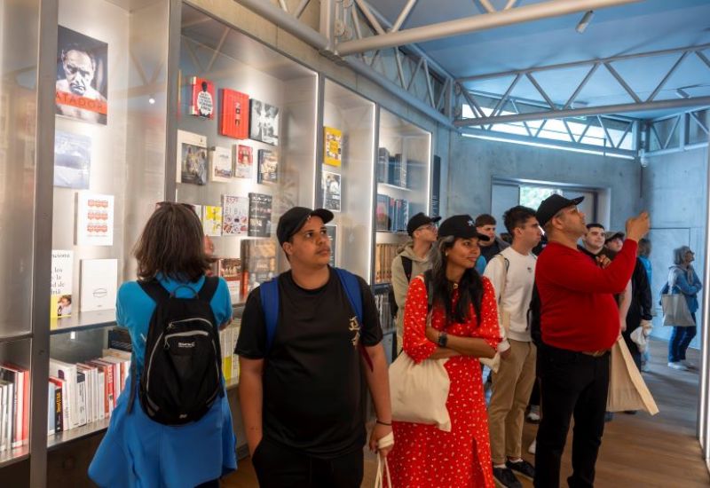 Interior de una exposici&oacute;n luminosa. Varias personas est&aacute;n de pie observando vitrinas con libros y objetos colocados cuidadosamente. Todo est&aacute; ordenado como en una biblioteca moderna.