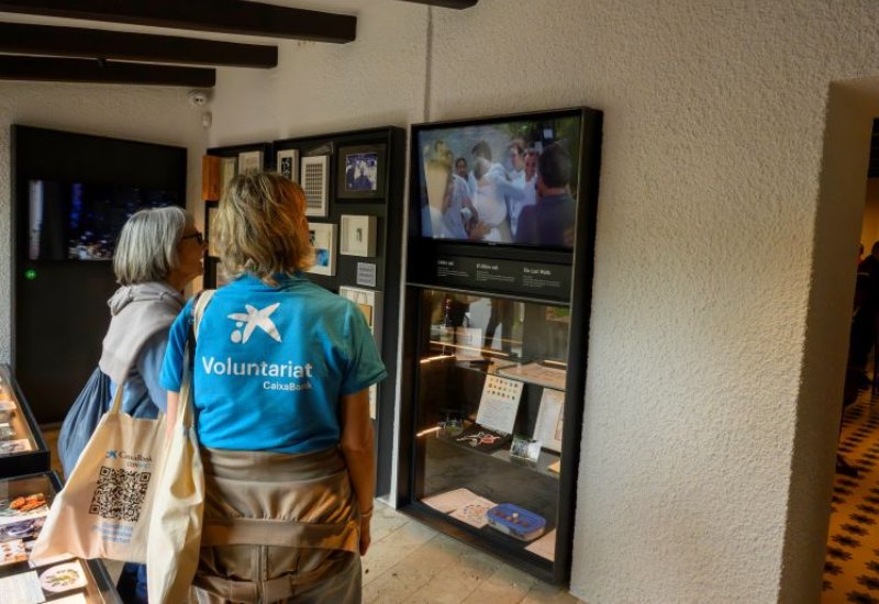 Voluntarias mirando una pantalla y vitrina durante la actividad. Interior del museo. Dos voluntarias observan una vitrina con objetos mientras delante hay una pantalla con un vídeo. El espacio es tranquilo, con cuadros y paneles informativos en las paredes.