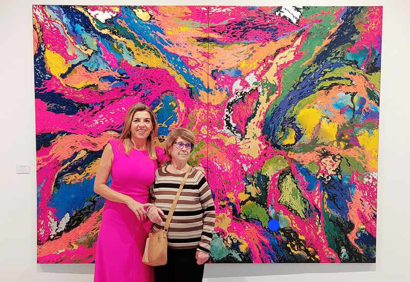 Una mujer voluntaria de CaixaBank posa, con un vestido rosa, delante de un cuadro abstracto junto a Luc&iacute;a, la anciana con la que comparte actividades