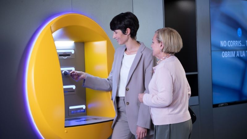 Empleada de CaixaBank atendiendo, en un cajero, a una mujer mayor.