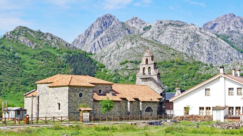 Burón: naturaleza y paisajes en la montaña de León Iglesia de piedra con campanario en Burón, rodeada de casas y prados verdes, con montañas rocosas al fondo bajo cielo despejado