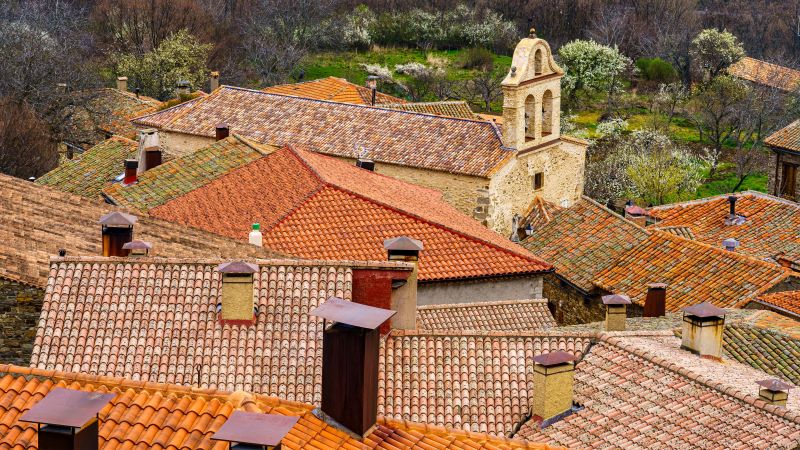 La Hiruela: pueblo con encanto en Madrid con arquitectura tradicional Tejados de teja rojiza en primer plano con chimeneas y una iglesia de piedra entre viviendas tradicionales, rodeadas de vegetación