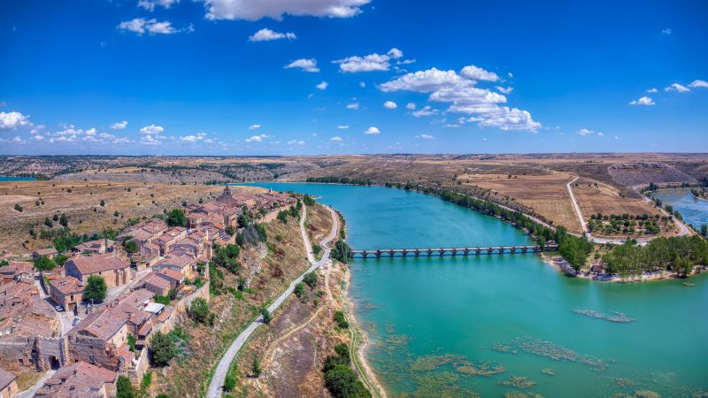 Maderuelo, un pueblo medieval con encanto en Segovia Vista aérea de Maderuelo con casas de piedra junto a un río turquesa curvado, un puente sobre el agua y paisaje seco bajo cielo azul con nubes
