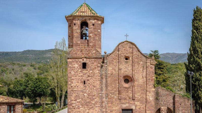 El Brull (Barcelona): arquitectura del medievo en un entorno privilegiado Iglesia de piedra en el Brull junto a la carretera, con campanario y edificio anexo, rodeada de árboles y montañas bajo cielo azul claro.
