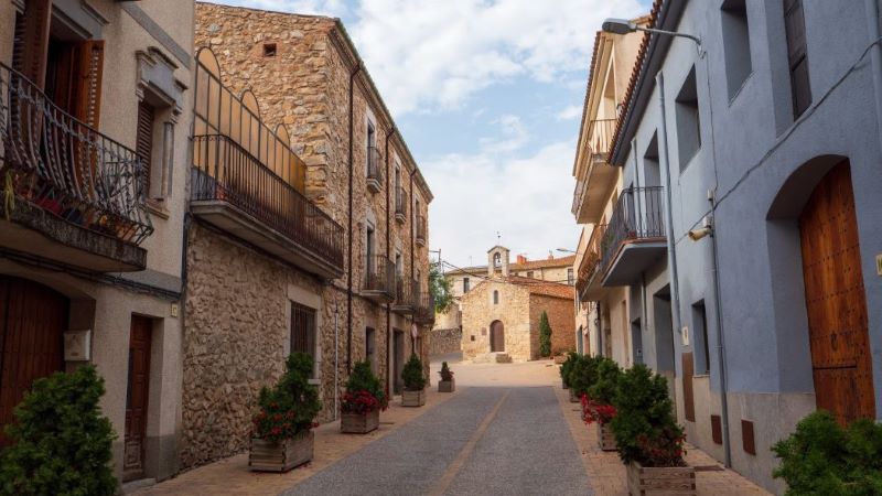 Capmany (Girona): Prehistoria entre viñedos Calle estrecha de Capmany con casas de piedra y balcones, macetas a los lados y una iglesia al fondo bajo cielo parcialmente nublado.