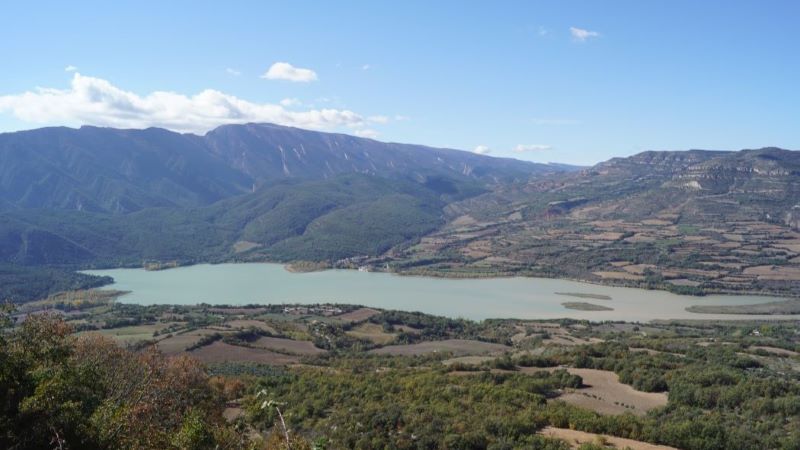 Llimiana (Lleida): montaña, río y embalse con vestigios del románico Vista panorámica del embalse rodeado de montañas y campos en Llimiana, con laderas verdes y cielo despejado, tomada desde un mirador elevado.