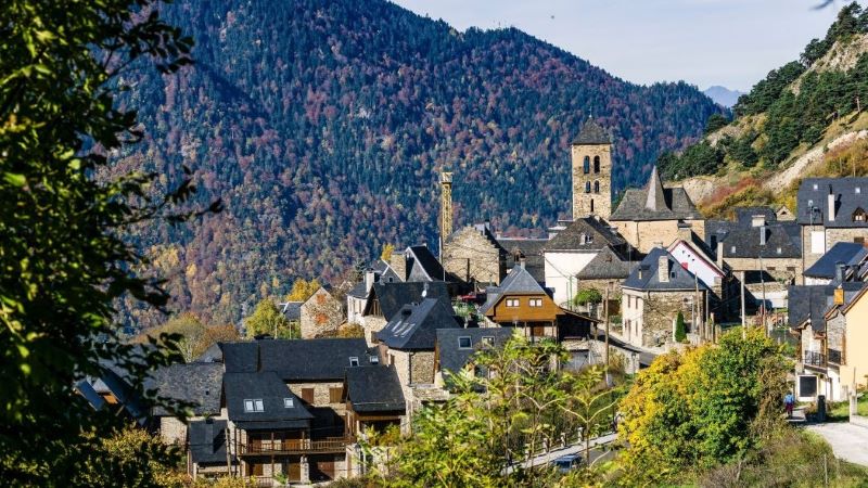 Vilamòs (Lleida): una inmersión en la tradición aranesa en plena Occitania Pueblo de montaña con casas de piedra y tejados oscuros en Vilamòs, con iglesia y torre, rodeado de bosques y montañas en otoño visto desde una ladera