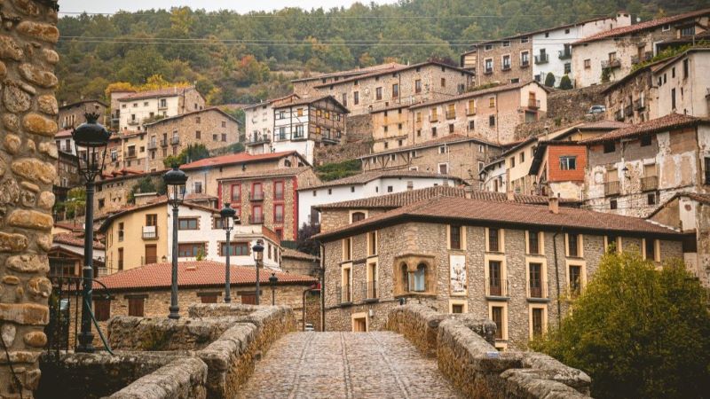 Villoslada de Cameros (La Rioja): huellas de otras culturas en un pueblo de tradición trashumante Puente de piedra que conduce a un pueblo de montaña con casas de piedra y tejados rojizos, en una ladera rodeada de bosque.